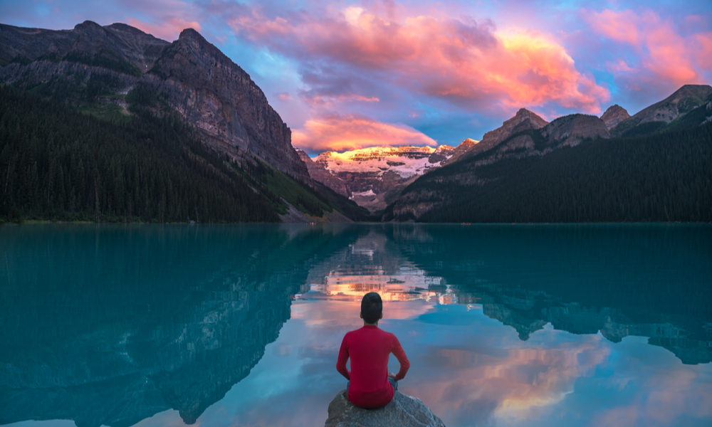 Top Domestic Destinations in Canada: A man in red sit on rock watching Lake Louise in the morning sun light with colorful clouds and reflections