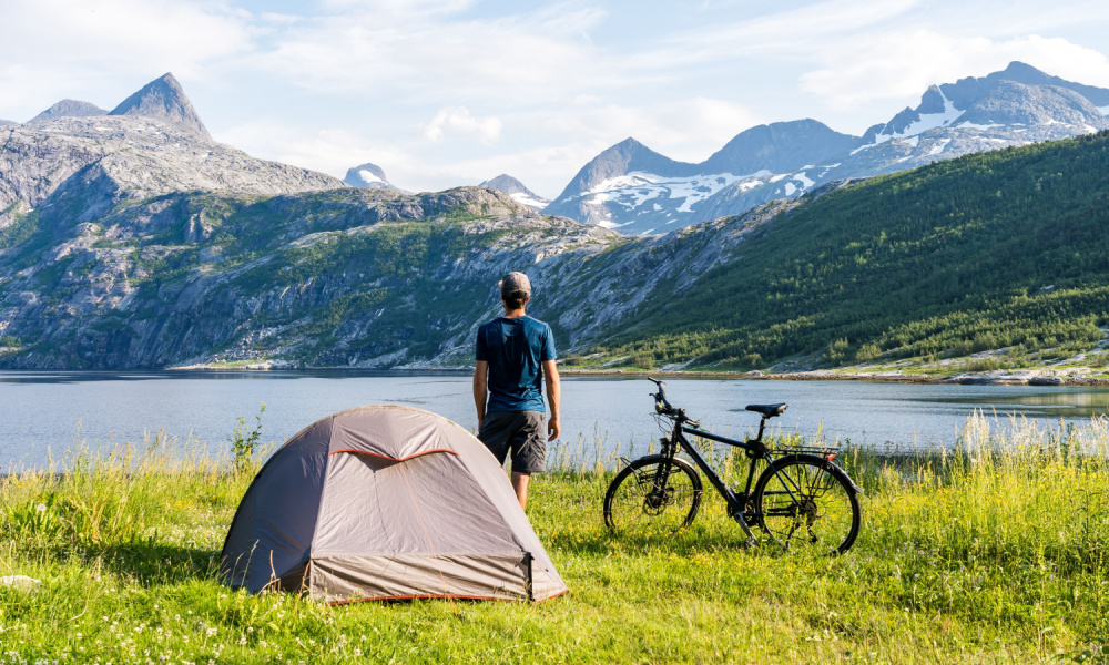 Man standing next to his trekking tent and bike on a bike travel through Norway with fjord and mountains in the background